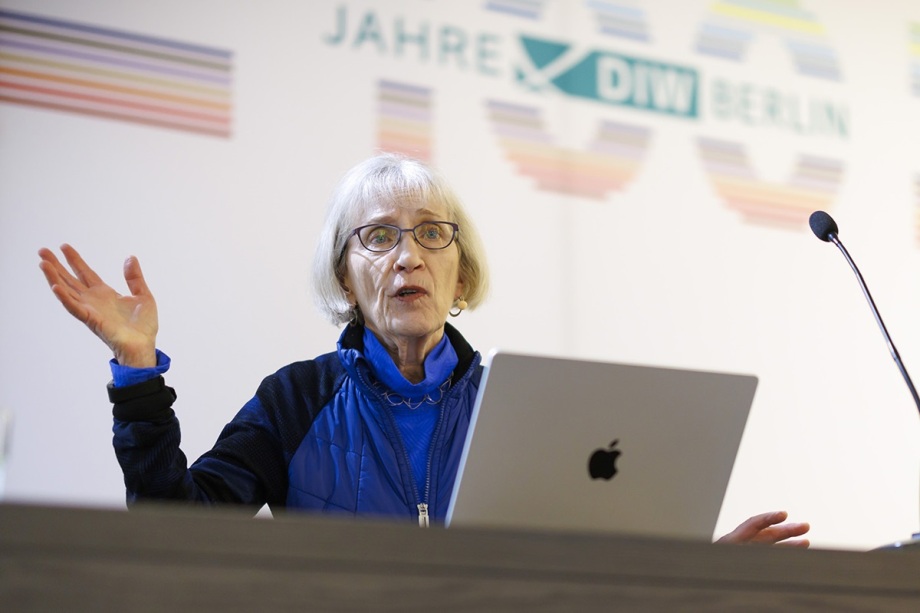 Von "Dads" und "Duds" - Claudia Goldin erklärte auch, wie Vaterschaft die Berufstätigkeit von Müttern beeinflusst.; Das Foto zeigt eine Person, die an einem Rednerpult steht und vor einem Laptop spricht. Die Frau ist Nobelpreisträgerin Claudia Goldin. Der Laptop ist silbern und trägt das Apple-Logo. Die Person trägt eine blaue Jake mit mehreren Schichten, darunter ein Oberteil. Beide Arme sind erhoben, die Hände sind geöffnet, als würde etwas betont oder erklärt. Rechts im Bild ist ein Mikrofon auf einem langen, schmalen Ständer zu sehen, das in Richtung der Person zeigt. Im Hintergrund befindet sich eine weiße Wand mit einem großen Schriftzug „100 Jahre DIW Berlin“. Die 100 Jahre bestehen aus farbigen Linien. Die Beleuchtung ist hell und gleichmäßig, was den Eindruck eines professionellen Vortrags vermittelt.