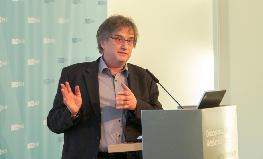 Jürgen Schupp bei der Veranstaltung im DIW Berlin, bei dem die finnischen Forscher*innen von ihrem Projekt berichteten.; Auf dem Foto ist ein Mann mittleren Alters zu sehen, der an einem Rednerpult steht und offenbar einen Vortrag hält. Er trägt ein dunkles Sakko über einem hellgrauen Hemd, das oben leicht geöffnet ist. Sein graues Haar ist mittellang und etwas zerzaust, er trägt eine Brille. Mit der rechten Hand gestikuliert er, während die linke Hand locker vor seinem Körper ruht. Vor ihm steht ein Mikrofon und ein aufgeklappter Laptop. Im Hintergrund ist eine hellgrüne Wand mit dem Logo „DIW Berlin“ zu sehen, was auf eine Veranstaltung des Deutschen Instituts für Wirtschaftsforschung hinweist. Der Mann wirkt konzentriert und engagiert, als wolle er einen wichtigen Punkt hervorheben.