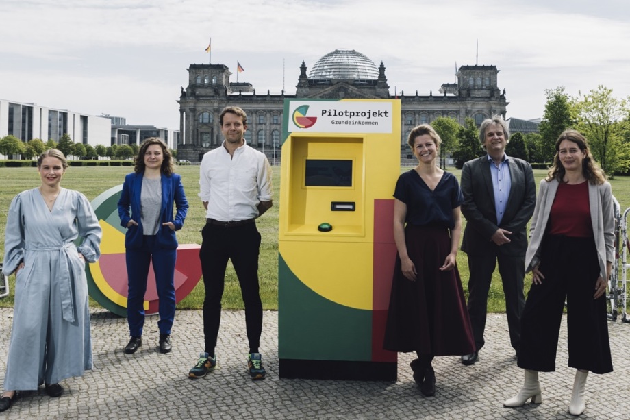 Zum Start des Pilotprojekts vor dem Bundestag: Janine Busch, Miriam Witz und Michael Bohmeyer vom Verein Mein Grundeinkommen e.V., Susann Fiedler von der Universität Wien, Jürgen Schupp vom DIW Berlin, und Maheba Goedeke Tort vom Verein Mein Grundeinkommen e.V. (v.l.n.r.); Das Bild zeigt sechs Personen, die im Freien vor einem gelb-grünen Kiosk stehen. Auf dem Kiosk steht die Aufschrift „Pilotprojekt Grundeinkommen“. Im Hintergrund ist ein großes historisches Gebäude mit einer Glaskuppel zu sehen, vermutlich das Reichstagsgebäude in Berlin. Die Personen tragen entweder Business- oder Freizeitkleidung und stehen locker um den Kiosk herum. Die Szene wirkt wie eine öffentliche Präsentation oder ein Auftakt zu einem gesellschaftlichen Projekt rund um das Thema Grundeinkommen.