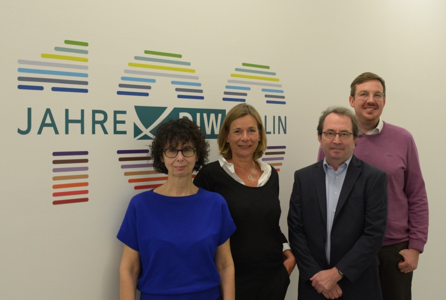 Die vier Chefredakteur*innen des Wochenberichtsredaktion vor dem 100 Jahre DIW-Logo. Kristina van Deuverden und Pio Baake (vorne) sind wissenschaftliche Chefredakteure, Claudia Cohnen-Beck und Sebastian Kollmann sind Teil der Kommunikationsabteilung. ; Vor einer Wand mit dem Schriftzug „100 JAHRE DIW BERLIN“ und einem bunten Design stehen vier Menschen. Ganz links steht eine Person mit Brille und einem blauen Oberteil. Daneben eine Person mit einem schwarzen Oberteil, das einen weißen Kragen hat. Die dritte Person trägt ebenfalls eine Brille und ist in einem dunklen Anzug mit einem hellen Hemd gekleidet. Ganz rechts steht eine Person mit Brille, einem lila Pullover und brauner Hose. Die Personen sind in zwei Reihen angeordnet – zwei vorne, zwei hinten – und blicken in Richtung Kamera, als wäre es ein offizielles Gruppenfoto.
