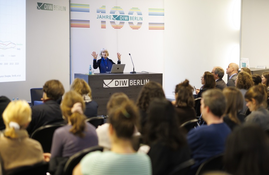 Nobelpreisträger Claudia Goldin war am 6.11.2025 zu Gast im DIW Berlin.; Das Foto zeigt einen Konferenzraum mit vielen sitzenden Personen, die alle nach vorne auf eine Bühne blicken. Auf der Bühne steht ein Rednerpult mit dem Logo „DIW Berlin“. Dahinter befindet sich eine weiße Wand mit dem Schriftzug „Jahre DIW Berlin“ und farbigen Linien, die dekorativ angeordnet sind. Auf dem Pult steht ein silberner Laptop mit Apple-Logo, daneben eine Wasserflasche und ein Mikrofon auf einem schmalen Ständer. Die Person am Pult hat beide Arme erhoben, als würde etwas betont oder erklärt. Die Person ist Nobelpreisträgerin Claudia Goldin. Links im Bild ist eine große Projektionsfläche zu sehen, auf der ein Diagramm mit Linien dargestellt ist. Der Raum ist hell beleuchtet, die Wände sind weiß, und die Sitzreihen sind dicht besetzt. Insgesamt vermittelt das Bild den Eindruck eines formellen Vortrags oder einer Präsentation vor einem konzentrierten Publikum.