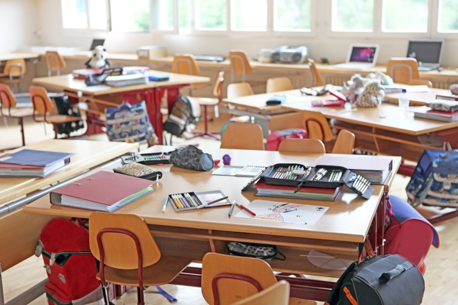 Auch bessere Bildungschancen zu ermöglichen, kann Kriminalitätsprävention sein.; Das Foto zeigt ein helles Klassenzimmer mit mehreren Gruppentischen, die jeweils von vier Stühlen umgeben sind. Auf den Tischen liegen verschiedene Schulsachen wie aufgeschlagene Ordner, Mäppchen, Stifte und Papier mit Zeichnungen. Unter den Tischen hängen bunte Schulranzen und Rucksäcke. Im Hintergrund stehen weitere Tische mit Computern und Laptops sowie einige Gegenstände wie Stofftiere und Taschen. Der Raum hat große Fenster, durch die Tageslicht hereinfällt, und der Boden besteht aus hellem Holz. Die Atmosphäre wirkt lebendig und ungeordnet, als wäre der Unterricht gerade unterbrochen oder die Schüler nicht im Raum.