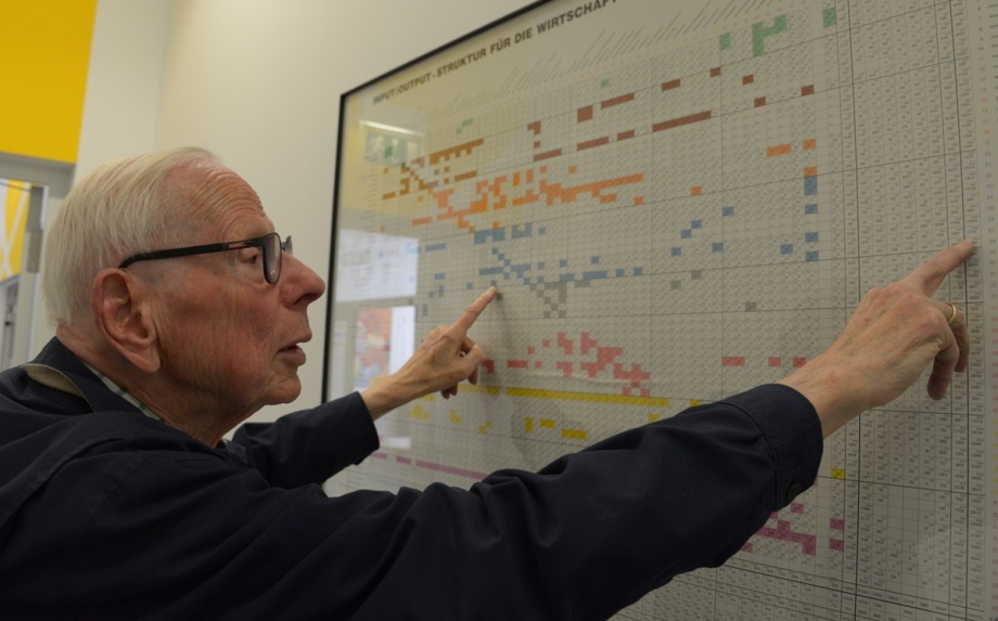 Reiner Stäglin erklärt „seine“ Input-Output-Tabelle als Wandtafel.; Auf dem Bild steht Reiner Stäglin vor einer großen, gerahmten Input-Output-Tabelle im DIW Berlin. Er zeigt mit beiden Händen auf verschiedene Stellen der Tabelle, die aus vielen kleinen, farbigen Kästchen und Zahlen besteht. Die Tabelle veranschaulicht wirtschaftliche Zusammenhänge zwischen verschiedenen Branchen. Im Hintergrund sind helle Wände und ein gelbes Wandstück zu sehen.