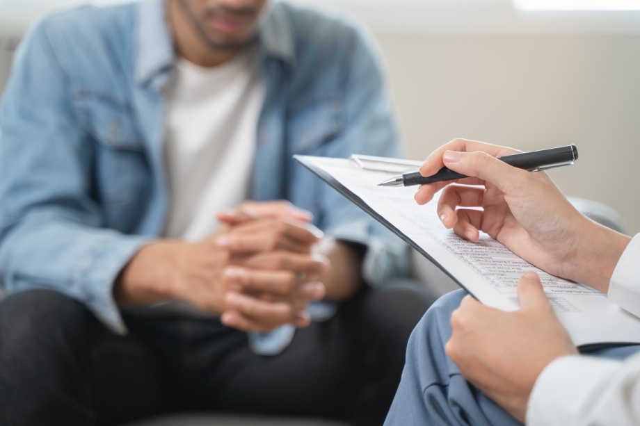 Hilfe nimmt eher in Anspruch, wer nicht aufgrund von psychischen Problemen stigmatisiert wird.; Das Foto zeigt eine Gesprächssituation in einem hellen Raum. Im Vordergrund ist eine Hand zu sehen, die einen Stift hält und auf ein Klemmbrett mit beschriebenem Papier zeigt. Die Schrift auf dem Papier ist nicht lesbar, aber es wirkt wie Notizen oder ein Formular. Im Hintergrund sitzt eine Person mit verschränkten Händen, die auf den Knien ruhen. Die Person trägt ein helles Oberteil und eine offene Jeansjacke. Der Fokus des Bildes liegt auf dem Klemmbrett und der Hand mit dem Stift, während die sitzende Person leicht unscharf dargestellt ist. Das Bild vermittelt den Eindruck eines Beratungsgesprächs oder einer professionellen Besprechung.