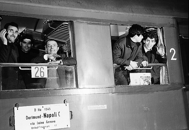 Das Bild zeigt eine Szene in einem Zugabteil. Mehrere Personen sitzen dicht nebeneinander auf gepolsterten Sitzbänken. Die Kleidung wirkt schlicht und praktisch, passend für eine Reise. Einige Personen haben Gepäckstücke bei sich, darunter Taschen und Koffer, die entweder auf dem Boden oder auf den Ablagen verstaut sind. Die Fenster des Zuges lassen Tageslicht herein, wodurch die Umgebung hell wirkt. Die Stimmung des Bildes vermittelt den Eindruck einer längeren Fahrt, möglicherweise eine Reise zu einem Arbeitsort. Das Foto ist in Schwarz-Weiß gehalten, was ihm einen historischen Charakter verleiht.