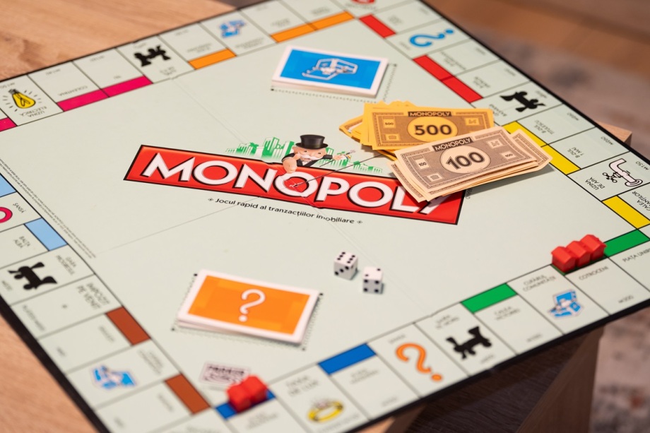 Monopoly – ein viel beliebtes Gesellschaftsspiel. Warum Monopole in der Realität ein Problem sind, erklärt Tomaso Duso im Interview.; Das Bild zeigt ein Monopoly-Spielbrett auf einem Tisch. In der Mitte prangt groß das rote „Monopoly“-Logo mit dem bekannten Maskottchen, dem Herrn mit Zylinder. Auf dem Spielfeld liegen verschiedene Spielelemente: Rechts oben befinden sich Spielgeldscheine in Gelb und Grau mit den Werten 500 und 100, unten links ist ein Stapel oranger Ereigniskarten mit einem Fragezeichen zu sehen. Davor liegen zwei Würfel, die eine neue Spielrunde andeuten. Am unteren Rand stehen mehrere rote Häuser, die als Spielfiguren oder Investitionen im Spiel dienen. Die Spielfelder am Rand zeigen die typischen Straßen, Bahnhöfe und Sonderfelder. Durch die gewählte Perspektive wirkt es so, als sei das Spiel gerade im Gange oder bereit für den nächsten Zug.