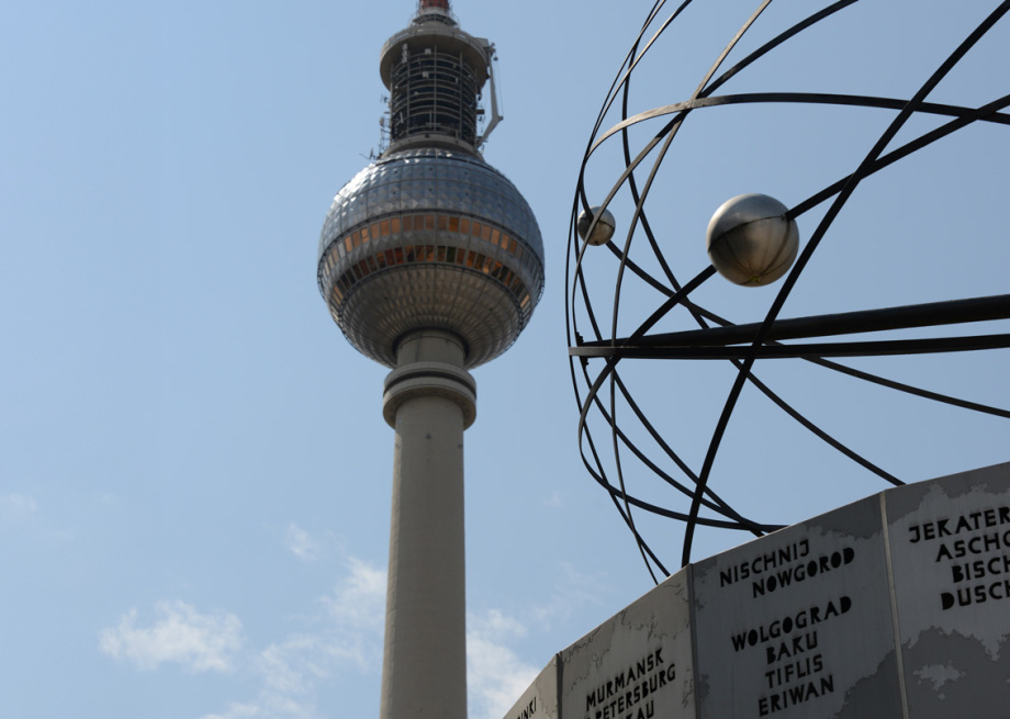 Das Bild zeigt den Berliner Fernsehturm und die Weltzeituhr auf dem Alexanderplatz in Berlin. Der Fernsehturm ist zentral im Hintergrund sichtbar und ragt mit seiner markanten Kugel und Antennenspitze hoch in den grauen, bewölkten Himmel. Im Vordergrund befindet sich die Weltzeituhr, ein kreisförmiges Kunstwerk mit Metallstreben, die ein stilisiertes Planetensystem darstellen. Darunter sind mehrere Tafeln mit Städtenamen und Zeitzonen angebracht. Das Bild vermittelt eine urbane, moderne Atmosphäre, verstärkt durch das wolkenverhangene Wetter.