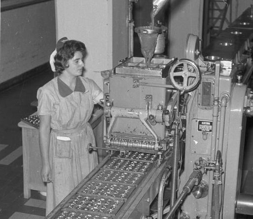 Eine Frau arbeitet 1962 am Fließband einer Schokoladenfabrik.; Das Foto zeigt eine historische Fabrikszene in Schwarz-Weiß. Im Vordergrund verläuft ein Förderband, auf dem zahlreiche kleine, runde Objekte in geordneten Reihen liegen, vermutlich Pralinen oder Schokoladenprodukte. Das Förderband führt zu einer großen, mechanischen Maschine mit mehreren Hebeln, Rohren und einem Trichter, aus dem Material eingefüllt wird. Die Maschine wirkt massiv und industriell, mit sichtbaren Metallteilen und einem großen Handrad an der Seite. Daneben steht eine Person in Arbeitskleidung, bestehend aus einem knielangen Kleid mit kurzem Ärmel und einer Schürze. Im Hintergrund sind weitere Teile der Produktionsanlage und ein Tisch zu erkennen. Die gesamte Szene vermittelt den Eindruck einer Schokoladenfabrik.
