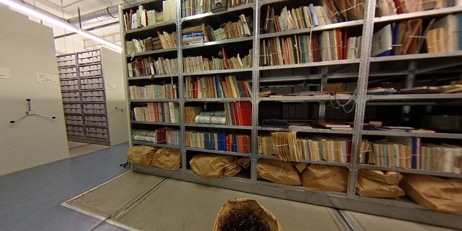 Im Stasi-Unterlagen-Archiv lässt sich wenig über das DIW Berlin finden.; Das Foto zeigt das Stasi-Unterlagen-Archiv mit hohen, metallenen Regalen, die bis zum Rand mit Akten und Dokumenten gefüllt sind. Die Akten sind unterschiedlich groß und in verschiedenen Farben gebunden, viele sind mit Schnüren oder Bändern zusammengebunden. Im Hintergrund befindet sich ein weiterer Satz von Regalen, die verschiebbar zu sein scheinen. Die Beleuchtung ist hell und funktional, typisch für ein Archiv. 