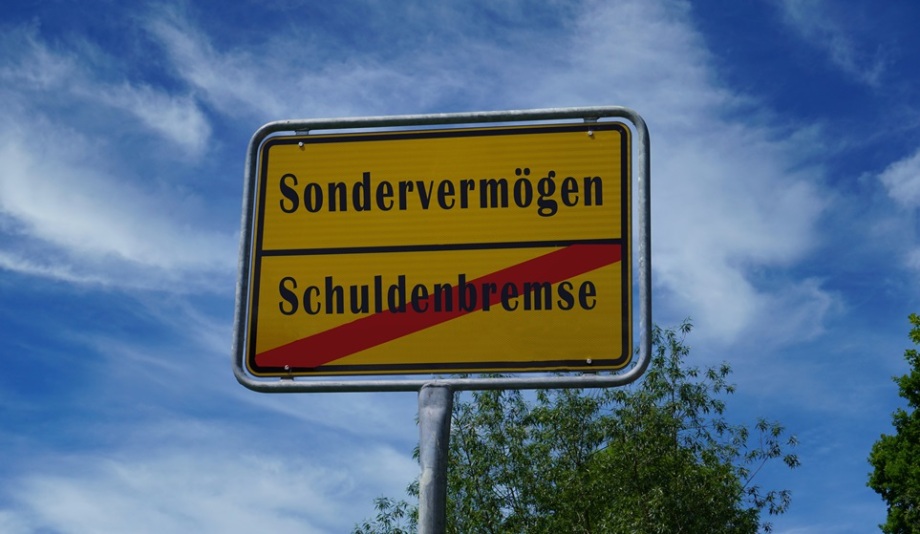 Mit einem Sondervermögen sollen Investitionen ermöglicht werden.; Das Bild zeigt ein gelbes Ortsschild mit schwarzem Rahmen, ähnlich einem offiziellen deutschen Verkehrsschild. Darauf steht oben das Wort "Sondervermögen" und darunter das Wort "Schuldenbremse", das von einem roten diagonalen Strich durchgestrichen ist. Der Hintergrund besteht aus einem blauen Himmel mit weißen Wolken sowie grünen Bäumen.