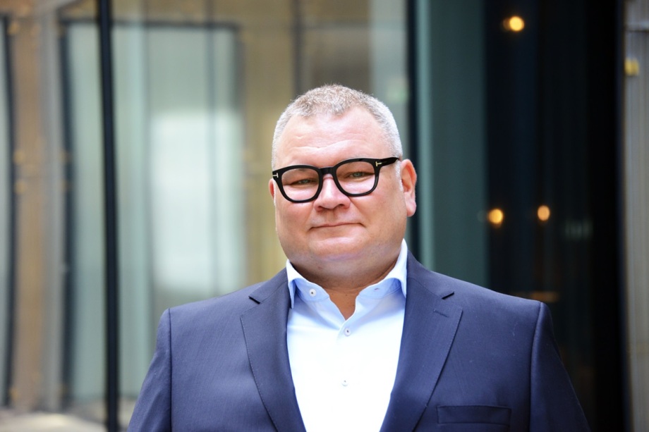 Lars Handrich ist Geschäftsführer der DIWEcon.; Das Bild zeigt einen Mann mittleren Alters in einem professionellen Umfeld. Er steht im Vordergrund, leicht lächelnd, mit direktem Blick in die Kamera. Der Mann hat kurzes, graues Haar und trägt eine markante, schwarze Brille mit dickem Rahmen. Er ist formell gekleidet, mit einem dunkelblauen Anzug und einem weißen Hemd ohne Krawatte.<br />
<br />
Der Hintergrund ist leicht unscharf und zeigt eine moderne Glasfassade mit reflektierenden Flächen, vermutlich der Eingang eines Bürogebäudes oder einer Institution.