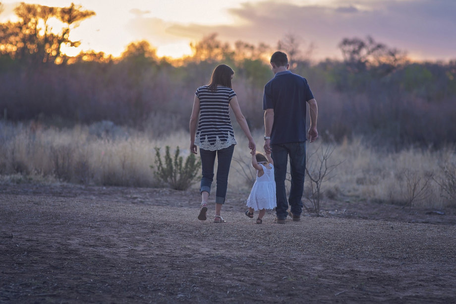 Bildtitel: Familie beim Sonnenuntergang<br />
Beschreibung: Das Bild zeigt eine Familie, bestehend aus zwei Erwachsenen und einem kleinen Kind, die auf einem Feldweg bei Sonnenuntergang spazieren gehen.<br />
Visuelle Elemente:<br />
Personen:<br />
Ein Erwachsener, vermutlich der Vater, hält die Hand des Kindes.<br />
Ein weiterer Erwachsener, vermutlich die Mutter, geht auf der anderen Seite des Kindes.<br />
Das Kind, das ein weißes Kleid trägt, wird von beiden Erwachsenen an den Händen gehalten.<br />
Umgebung: Der Weg, auf dem sie gehen, ist von trockenem Gras und Büschen umgeben.<br />
Im Hintergrund ist ein Sonnenuntergang zu sehen, der den Himmel in warmen Farben wie Orange und Gelb taucht.<br />
Die Landschaft wirkt ruhig und friedlich, mit sanften Hügeln und spärlicher Vegetation.<br />
Zusätzliche Informationen: Das Bild vermittelt ein Gefühl von Zusammengehörigkeit und Familienzeit in einer natürlichen Umgebung.<br />
Die Kleidung der Personen deutet auf eine entspannte, informelle Atmosphäre hin.<br />
Diese Beschreibung bietet einen Überblick über die Szene einer Familie, die gemeinsam einen Spaziergang bei Sonnenuntergang genießt.