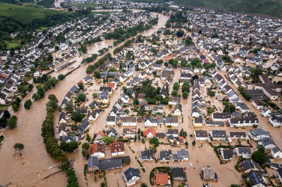 Die verheerenden Schäden der Flut im Ahrtal.; Das Bild zeigt eine Luftaufnahme einer stark überschwemmten Stadt. Zahlreiche Straßen und Häuser stehen tief im braunen, schlammigen Hochwasser, das vermutlich von einem Fluss stammt, der sich über sein Ufer ergossen hat. Das Wasser zieht sich wie breite, verzweigte Bänder durch die Stadt und umschließt ganze Straßenzüge. Viele Dächer von Ein- und Mehrfamilienhäusern ragen aus den Fluten, während Gärten, Höfe und Straßen komplett unter Wasser stehen. Die Häuser sind größtenteils in hellen Farben gestrichen, mit dunklen Dächern, und liegen dicht beieinander. Vereinzelt sieht man Bäume, deren Kronen aus dem Wasser ragen. Im Hintergrund setzen sich die überschwemmten Bereiche weiter fort, bis zu den angrenzenden grünen Hügeln. Das Ausmaß der Katastrophe ist deutlich sichtbar – die Wassermassen haben große Teile des Ortes vollständig lahmgelegt.