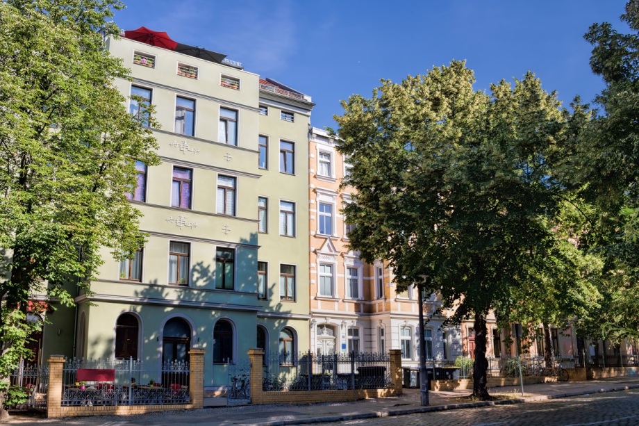 Viele Geflüchtete gaben an, bei der Wohnungssuche Diskriminierung erlebt zu haben.; Das Foto zeigt eine Reihe mehrstöckiger Wohnhäuser in einer städtischen Umgebung. Die Gebäude sind modern und in hellen, neutralen Farben wie Weiß und Beige gehalten. Sie haben große Fenster und Balkone, die teilweise mit Geländern versehen sind. Im Vordergrund ist ein Gehweg oder eine Straße zu erkennen, die von Bäumen und Sträuchern gesäumt wird, was dem Bild einen gepflegten und geordneten Eindruck verleiht. Der Himmel ist klar und blau, wodurch die Szene hell und freundlich wirkt. Insgesamt vermittelt das Bild den Eindruck eines ruhigen Wohnviertels mit zeitgemäßer Architektur.