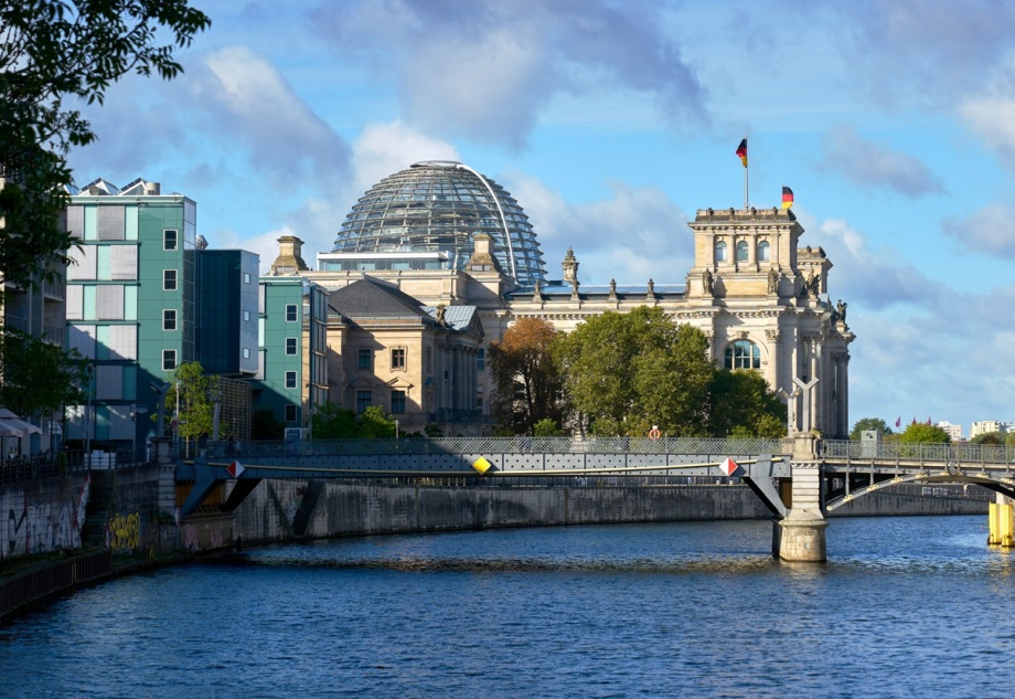 Dazu, dass Geflüchtete sich in Deutschland weniger willkommen fühlen, trägt auch der politische Diskurs bei.; Das Foto zeigt die gläserne Kuppel des Reichstagsgebäudes in Berlin, die zentral im oberen Bereich des Bildes zu sehen ist. Darunter erstreckt sich die historische Fassade des Reichstags mit heller Sandsteinstruktur und klassischen Architekturdetails. Mehrere deutsche Flaggen sind auf dem Dach des Gebäudes erkennbar. Im Vordergrund fließt ein breiter Fluss, dessen Wasseroberfläche das Licht reflektiert. Über den Fluss führt eine moderne Fußgängerbrücke mit geschwungenem Design, die von links nach rechts verläuft. Am Ufer sind Bäume und Grünflächen sichtbar, die die Szene rahmen. Der Himmel ist klar und blau, mit wenigen weißen Wolken, was dem Bild eine helle und offene Stimmung verleiht. 