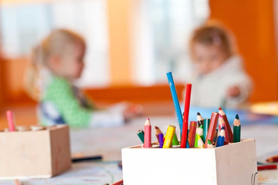 Das Foto zeigt eine Nahaufnahme eines weißen Behälters, der mit vielen bunten Buntstiften gefüllt ist. Die Stifte sind unterschiedlich lang und in kräftigen Farben wie Rot, Blau, Grün und Gelb. Der Behälter steht auf einem Tisch, der mit Papier bedeckt ist, auf dem Zeichnungen oder Kritzeleien zu erkennen sind. Im Hintergrund ist ein heller Raum mit warmen orangefarbenen Wänden zu sehen. Dort sitzen zwei Kinder an demselben Tisch, leicht unscharf dargestellt, offenbar beschäftigt mit Mal- oder Bastelarbeiten. Das Licht im Raum wirkt freundlich und hell, vermutlich durch große Fenster, die für eine angenehme Atmosphäre sorgen.