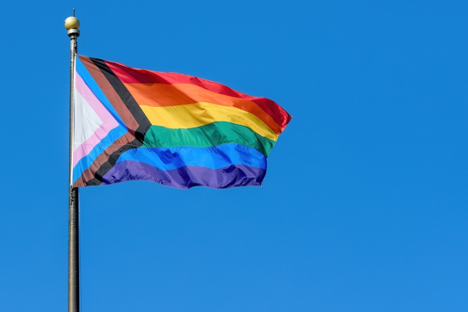 Wie sind die Lebenslagen und Diskriminierungserfahrungen queerer Menschen? Dazu forschte das DIW Berlin.; Auf dem Foto weht eine sogenannte Progress-Pride-Flagge an einem hohen Fahnenmast vor wolkenlosem, kräftig blauem Himmel. Das Tuch ist vom Wind aufgebläht, sodass seine Farben klar zu erkennen sind und schräg nach rechts auswehen. Im rechten, größeren Teil der Flagge verlaufen waagerecht die klassischen Regenbogenstreifen in Rot, Orange, Gelb, Grün, Blau und Violett. Am linken Rand ist ein nach rechts zeigendes, dreieckiges Muster zu sehen: außen ein schwarzer und ein brauner Streifen, die für Schwarze und andere People of Color sowie für Menschen stehen, die von Rassismus betroffen sind, und innen die Pastellfarben Rosa, Hellblau und Weiß der Transgender-Flagge. Oben auf dem dunklen Metallmast sitzt eine kleine, goldfarbene Kugel, die sich klar vom Himmel abhebt.