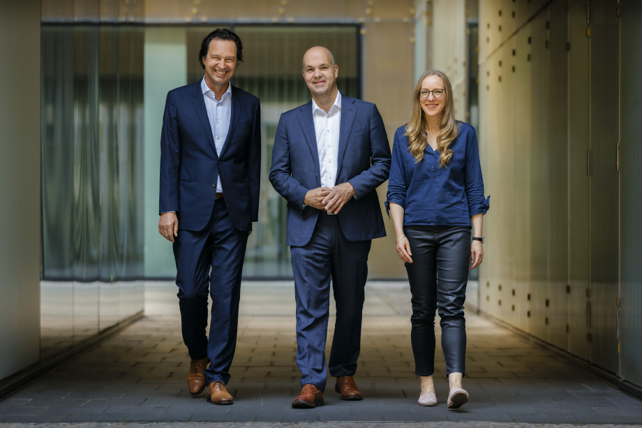 Der Vorstand des DIW Berlin (v.l.n.r.) Alexander S. Kritikos, Marcel Fratzscher, Denise Rüttinger; Drei Personen in Geschäftskleidung stehen nebeneinander in einem modernen Gebäude mit Glaswänden im Hintergrund. Zwei Männer und eine Frau lächeln und wirken professionell. Der Mann in der Mitte hat die Hände vor dem Körper gefaltet, während die anderen beiden die Arme locker an den Seiten halten. Der Hintergrund zeigt eine helle, moderne Architektur mit großen Fenstern und einer Tür.