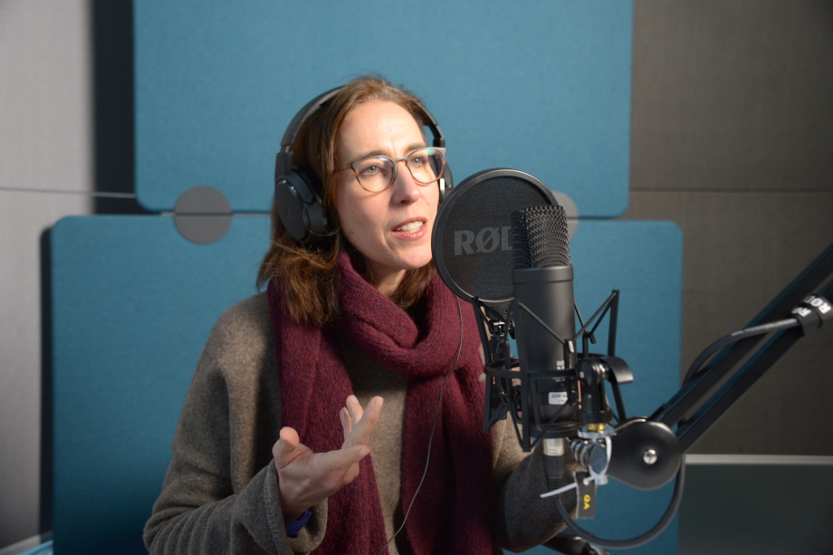 Katharina Wrohlich zu Gast im Podcast "Wirtschaft bewegt - 100 Jahre DIW Berlin".; Das Bild zeigt eine Frau, Katharina Wrohlich, in einem Tonstudio. Sie sitzt vor einem großen, professionellen Mikrofon mit einem Popfilter, auf dem das Markenlogo „RØDE“ zu sehen ist. Sie trägt große schwarze Kopfhörer über ihren braunen, schulterlangen Haaren und hat eine Brille mit rundlichen Gläsern. Sie ist in ein dunkelgraues Oberteil gekleidet und trägt dazu einen weinroten Schal. Ihre Hände sind in einer sprechenden Geste erhoben – die linke Hand ist geöffnet, die rechte nur teilweise im Bild sichtbar. Ihr Gesichtsausdruck ist engagiert, aufmerksam und freundlich – sie scheint gerade zu sprechen oder etwas zu erklären.<br />
<br />
Der Hintergrund besteht aus schallisolierenden Wandpaneelen in verschiedenen Blau- und Grautönen, wie sie in Tonstudios zur Verbesserung der Akustik üblich sind. Rechts im Bild ist ein schwarzer Mikrofonarm zu sehen, der das Mikrofon hält.
