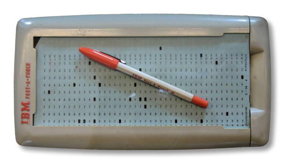 Handlocher und Lochkarte der International Business Machines Corporation (IBM).; Das Bild zeigt ein rechteckiges, graues Gerät namens „IBM Port-A-Punch“, auf dessen Oberfläche eine Lochkarte eingelegt ist. Die Lochkarte ist mit Zahlen und kleinen Feldern bedruckt, in die Löcher gestanzt werden können. Quer über das Gerät liegt ein Kugelschreiber mit roter Kappe und Spitze. Auf dem Stift steht ebenfalls „IBM Port-A-Punch“. Das Gerät und die Karte wirken technisch und stammen aus der Frühzeit der Computertechnik, als Daten noch auf Papier-Lochkarten gespeichert wurden.