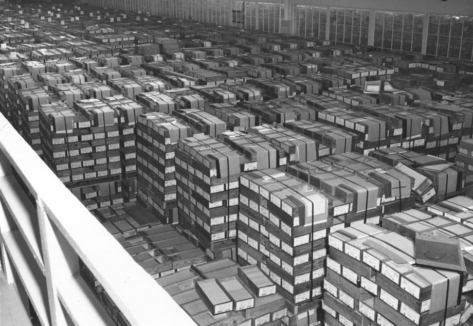 Lochkarten-Lager. Für einen Megabyte an Daten benötigt man etwa 12.500 Lochkarten.; Das Bild zeigt eine große Lagerhalle, die vollständig mit stapelweise aufgestellten Kartons gefüllt ist. Die Kartons sind in langen, geraden Reihen angeordnet und reichen bis tief in den Hintergrund des Raumes. Jeder Karton ist mit Bändern zusammengebunden, und auf den Stirnseiten sind Etiketten sichtbar. In diesen Kartons befinden sich Lochkarten, die früher zur Datenspeicherung und -verarbeitung in Computern genutzt wurden. Die gleichförmigen, dicht an dicht gestapelten Kartons vermitteln ein Bild von enormen Datenmengen, die hier physisch gelagert werden.