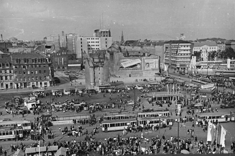 Eine Stadt im Wiederaufbau: Das Foto zeigt den Alexanderplatz 1951 – eine Zeit, als auch das DIW mit Ferdinand Friedensburg an einem Neubeginn arbeitete.; Das Bild zeigt den Berliner Alexanderplatz im Jahr 1951. Im Vordergrund sind zahlreiche Menschen zu sehen, die sich auf dem Platz bewegen. Straßenbahnen fahren und halten an verschiedenen Stellen. Im Hintergrund sind zerstörte Gebäude und Ruinen zu erkennen, die an die Folgen des Zweiten Weltkriegs erinnern. Ein markantes Bauwerk in der Bildmitte ist ein beschädigter Turm, umgeben von Werbeplakaten.