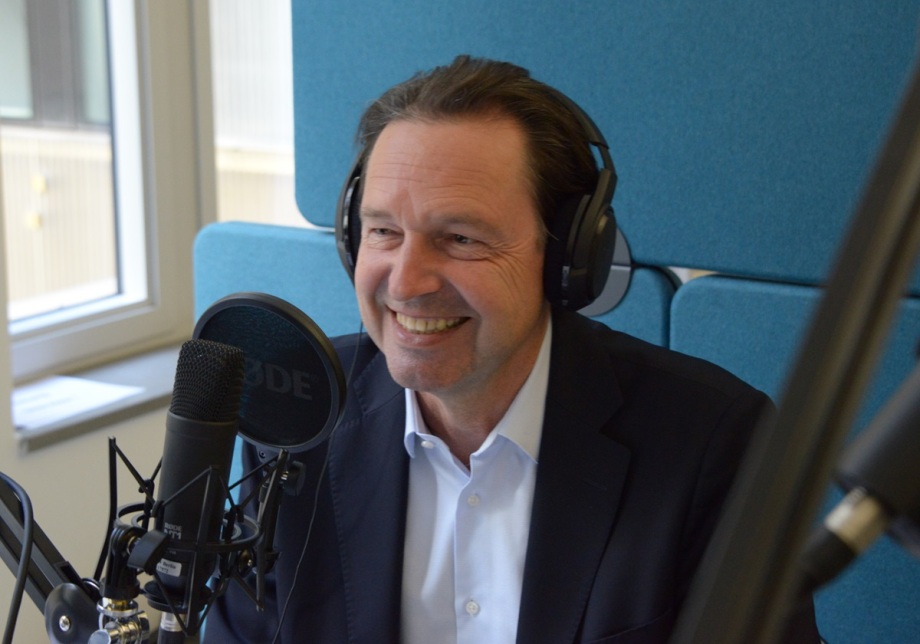 Alexander S. Kritikos, Mitglied des DIW-Vorstands, im Podcastgespräch; Alexander S. Kritikos, Mitglied des DIW-Vorstands, sitzt lächelnd in einem Studio und trägt ein dunkles Sakko über einem weißen Hemd. Er hat Kopfhörer auf und spricht in ein Mikrofon, das sich direkt vor ihm befindet. Im Hintergrund sieht man ein Fenster mit Tageslicht und einen Teil der Studioausstattung. Die Wände des Studios sind blau.