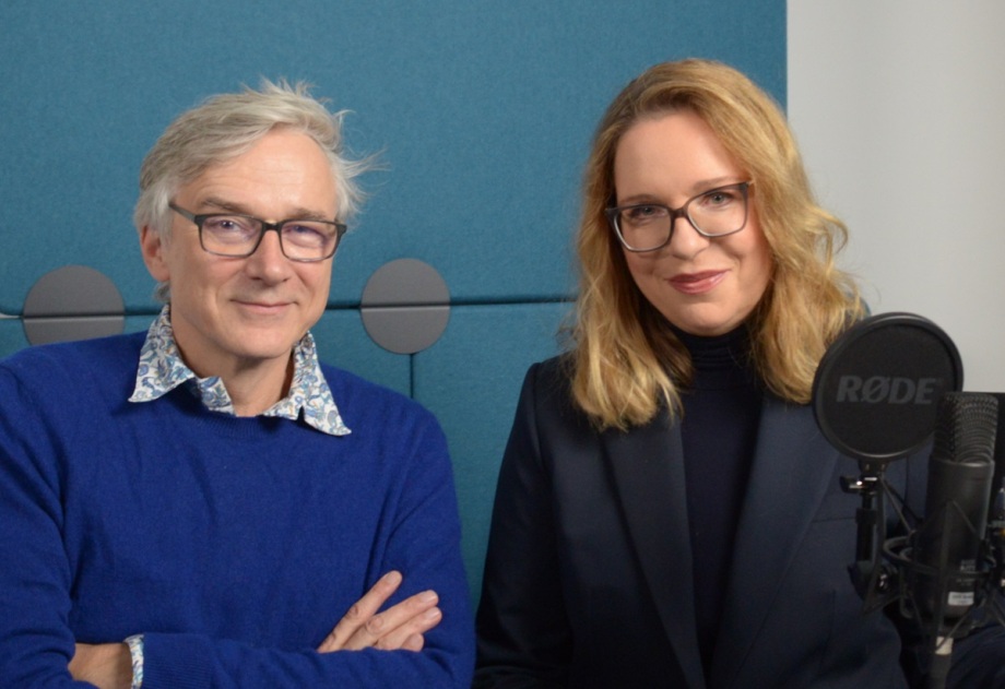 Podcast-Host Erich Wittenberg und Claudia Kemfert.; Das Foto zeigt zwei Personen, die nebeneinander vor einer blauen, gepolsterten Wand sitzen oder stehen. Beide tragen dunkle Kleidung: Die linke Person hat einen leuchtend blauen Pullover über einem Hemd mit auffälligem, gemustertem Kragen, die Arme sind vor der Brust verschränkt. Die rechte Person trägt ein dunkles Jackett über einem dunklen Oberteil, die Haare sind schulterlang und leicht gewellt. Rechts im Bild befindet sich ein schwarzes Mikrofon mit dem Markenaufdruck „RODE“, montiert auf einem Ständer, was auf eine Podcast- oder Interview-Situation hinweist. Die Umgebung wirkt schlicht und modern, mit Fokus auf die beiden Personen und das Mikrofon.