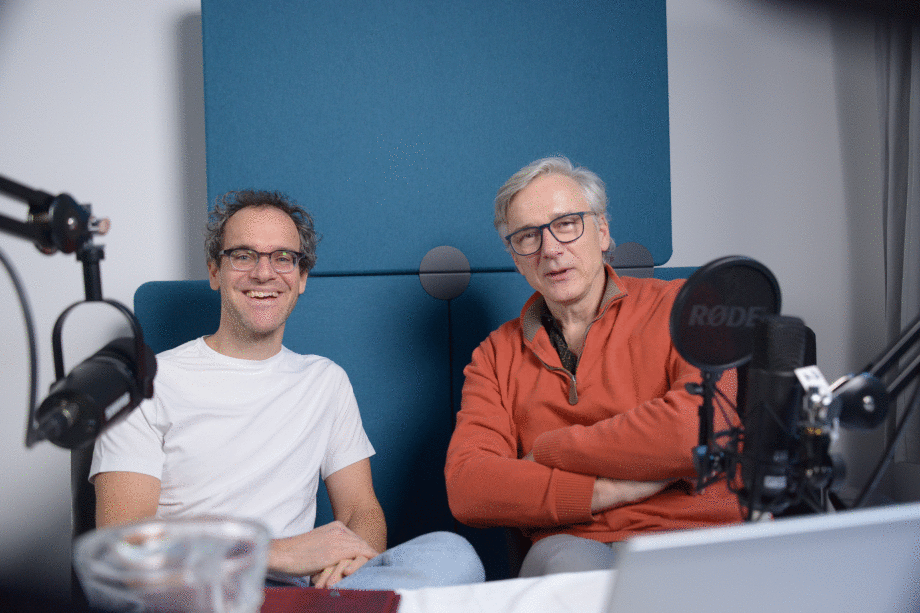 Wolf-Peter Schill (links) und Erich Wittenberg bei der Aufzeichnung der Podcastfolge zum Thema Wissenschaftskommunikation. ; Das Foto zeigt eine Szene in einem Aufnahmestudio für Podcasts. Zwei Personen sitzen sich an einem rechteckigen Tisch gegenüber. Auf dem Tisch liegt ein weißes Tuch, darauf stehen ein aufgeklappter Laptop, mehrere Blätter Papier, ein rotes Schreibheft, ein Smartphone sowie ein Glas und eine Glaskaraffe mit Wasser. Beide Personen tragen große Kopfhörer und sprechen in professionelle Mikrofone, die an beweglichen Halterungen befestigt sind. Die Mikrofone sind mit Pop-Schutz ausgestattet. Im Hintergrund ist ein großes Fenster zu sehen, durch das ein Gebäude mit Glasfassade erkennbar ist. Die Beleuchtung ist hell, und eine runde Studiolampe mit Diffusor sorgt für zusätzliches Licht. Die Atmosphäre wirkt technisch und konzentriert, typisch für eine Podcast-Produktion. 