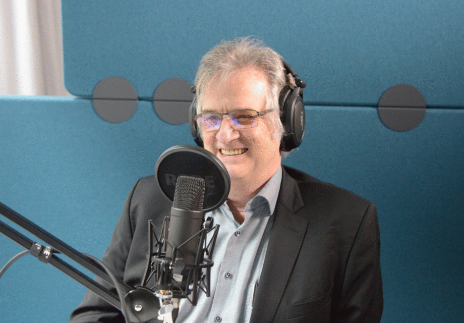 Jürgen Schupp bei der Podcastaufzeichnung zur Glücksforschung.; Das Foto zeigt eine Person, die in einem Tonstudio sitzt und an einer Aufnahme teilnimmt. Die Person trägt große, geschlossene Kopfhörer und sitzt vor einem professionellen Mikrofon, das auf einem verstellbaren Arm montiert ist. Das Mikrofon hat einen runden Popschutz mit der Aufschrift „RØDE“. Die Person trägt ein dunkles Sakko über einem hellblauen Hemd, das leicht geöffnet ist. Im Hintergrund ist eine schallisolierende Wand in einem kräftigen Blauton zu sehen, die mit runden, dunklen Elementen versehen ist. Die Beleuchtung wirkt gleichmäßig und hell, was auf eine kontrollierte Studio-Umgebung hinweist.