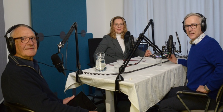 Im Gespräch mit Moderator Erich Wittenberg (rechts): Jürgen Blazejczak (links) und Claudia Kemfert (mitte).; Das Bild zeigt drei Personen, die an einem Tisch in einem Tonstudio sitzen und offenbar eine Podcast-Aufnahme machen. Alle tragen große Kopfhörer und sprechen in Mikrofone, die an beweglichen Halterungen befestigt sind. In der Mitte sitzt eine Frau mit blonden Haaren und Brille, Claudia Kemfert, sie trägt einen grauen Blazer und ein weißes Oberteil. Links sitzt ein älterer Mann, Jürgen Blazeczak, mit Brille und dunklem Pullover, rechts ein Mann mit grauem Haar, Brille und blauem Pullover über einem gemusterten Hemd, Erich Wittenberg. Auf dem Tisch liegen einige Papiere, Gläser mit Wasser und ein Wasserkrug. Der Raum wirkt ruhig und professionell eingerichtet, mit Schallschutzpaneelen an den Wänden.