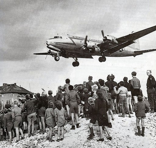 Auf diesem Foto stehen deutsche Kinder auf einem Schuttberg und beobachten ein US-Flugzeug, das über sie hinwegfliegt.; 