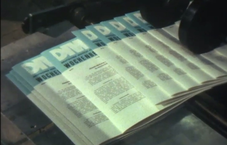 Der DIW-Wochenbericht kam 1980 aus der Druckmaschine in Berlin-Kreuzberg.; Dieses Bild zeigt eine Szene aus einer Druckerei. Mehrere frisch gedruckte Exemplare einer Publikation mit dem Titel „DIW Wochenbericht“ liegen nebeneinander auf einem Förderband oder einer Ablagefläche, offenbar gerade aus einer Druckmaschine kommend.<br />
<br />
Die Titelseiten der Hefte sind überwiegend hell (weiß oder cremefarben) mit einem blauen oberen Rand. Groß und fettgedruckt steht „DIW“ ganz oben, darunter das Wort „Wochenbericht“ in Großbuchstaben.<br />
<br />
Die Seiten sind dicht mit Text bedruckt, ohne Bilder oder auffällige Grafiken. Zwei große schwarze Walzen der Druckmaschine sind oben rechts im Bild zu sehen, was darauf hinweist, dass der Druckvorgang gerade stattfindet oder kurz davor abgeschlossen wurde.