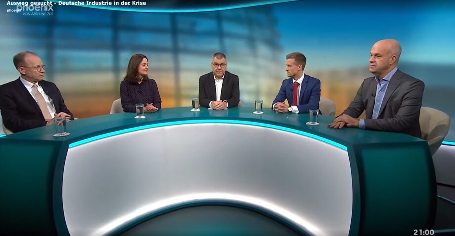 Bildtitel: Diskussionsrunde im Fernsehstudio zum Thema "Deutsche Industrie in der Krise" - Phoenixrunde vom 06.11.2025<br />
Beschreibung: Das Bild zeigt eine Diskussionsrunde in einem Fernsehstudio. Fünf Personen sitzen an einem halbkreisförmigen Tisch und diskutieren über das Thema "Deutsche Industrie in der Krise". Ganz rechts im Bild sitzt Marcel Fratzscher, Präsident des Deutschen Instituts für Wirtschaftsforschung. Die Sendung wird von Phoenix, einem gemeinsamen Sender von ARD und ZDF, ausgestrahlt.<br />
Visuelle Elemente:<br />
Hintergrund:<br />
Der Hintergrund zeigt ein Studio mit einer bläulichen Beleuchtung und einem leicht unscharfen Bild von Industrieanlagen oder Strommasten, was den thematischen Fokus der Diskussion unterstreicht.<br />
Oben links im Bild ist das Logo von Phoenix mit dem Schriftzug "phoenix von ARD und ZDF" zu sehen.<br />
Über dem Tisch steht der Titel der Sendung: "Ausweg gesucht - Deutsche Industrie in der Krise".<br />
Tisch und Teilnehmer: Der Tisch ist halbkreisförmig und in einem hellen Türkis-Ton gehalten. Auf dem Tisch stehen Wassergläser vor jedem Teilnehmer.<br />
Vier Personen sitzen an dem Tisch:<br />
Person 1 (links): Ein älterer Mann mit Brille und Anzug.<br />
Person 2: Eine Frau mit dunklen Haaren, die ein dunkles, gemustertes Oberteil trägt.<br />
Person 3: Ein älterer Mann mit Brille, graumeliertem Haar und Anzug.<br />
Person 4: Ein jüngerer Mann mit kurz geschnittenen Haaren, der einen dunklen Anzug mit Krawatte trägt.<br />
Person 5 (rechts): Ein glatzköpfiger Mann in einem dunklen Anzug mit offenem Hemdkragen.<br />
Körpersprache: Die Teilnehmer wirken aufmerksam und sind in die Diskussion vertieft. Einige haben die Hände gefaltet oder liegen auf dem Tisch, was eine entspannte, aber konzentrierte Atmosphäre vermittelt.<br />
Uhrzeit: Unten rechts im Bild ist die Uhrzeit "21:00" zu sehen, was darauf hinweist, dass die Sendung abends ausgestrahlt wird.<br />
Zusätzliche Informationen: Die Bildkomposition zielt darauf ab, eine professionelle und seriöse Diskussionsrunde darzustellen, die sich mit einem aktuellen und relevanten Thema der deutschen Industrie beschäftigt.<br />
Die visuelle Gestaltung des Studios und die Körpersprache der Teilnehmer vermitteln eine sachliche und fokussierte Atmosphäre.
