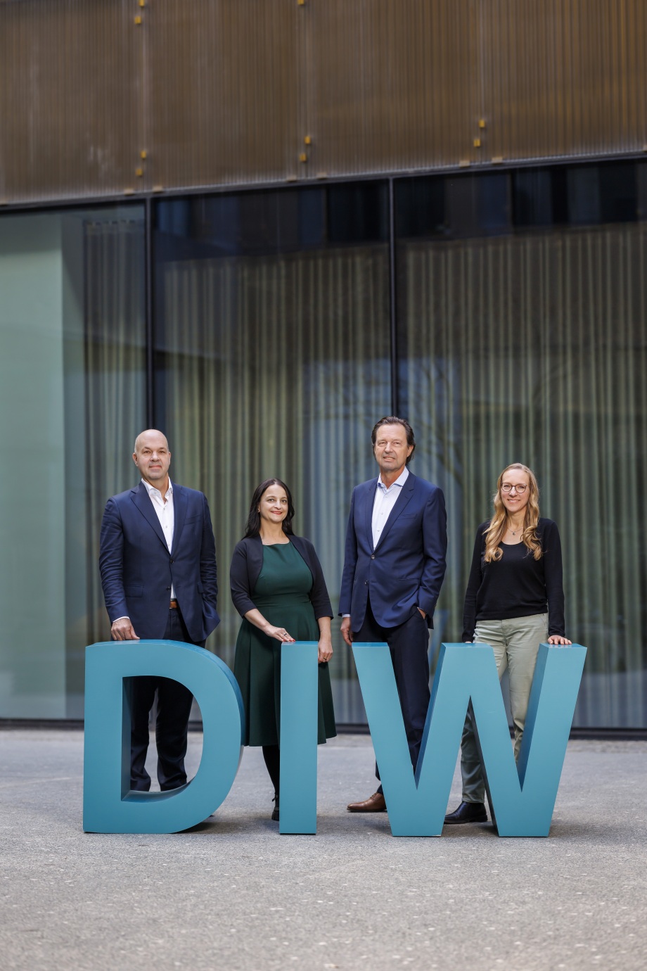 Der Vorstand des DIW Berlin (v.l.n.r.) Marcel Fratzscher, Sabine Zinn, Alexander S. Kritikos, Denise Rüttinger; Vier Personen in Geschäftskleidung stehen nebeneinander vor einem modernen Gebäude mit Glaswänden im Hintergrund. Vor ihnen stehen große Aufsteller der Buchstaben "DIW". Zwei Männer und zwei Frauen lächeln und wirken professionell. Der Hintergrund zeigt eine helle, moderne Architektur mit großen Fenstern.